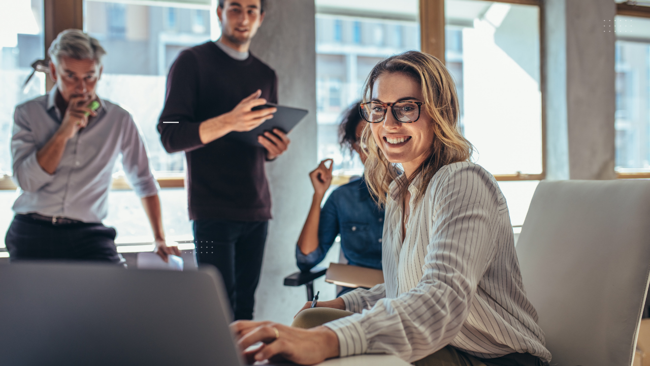Group of professionals in a modern office setting, collaborating on a business project. A smiling woman in glasses sits at the forefront working on a laptop, while three colleagues engage in discussion behind her. The image represents teamwork and a positive work environment, aligning with the blog post on building a successful Customer Experience (CX) strategy.
